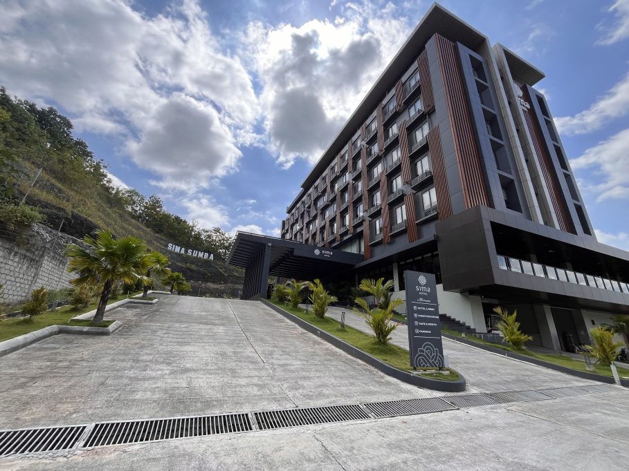 Sima Sumba Hotel, a modern multi-story building with dark grey and brown accents, stands on a green hillside with a paved entrance and palm trees.
