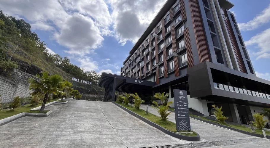 Sima Sumba Hotel, a modern multi-story building with dark grey and brown accents, stands on a green hillside with a paved entrance and palm trees.