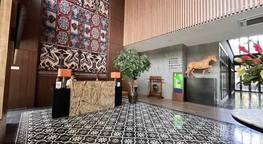 A luxurious hotel lobby features a large Sumba ikat tapestry, a reception desk, a potted plant, and a wooden horse sculpture on patterned black and white tiles.