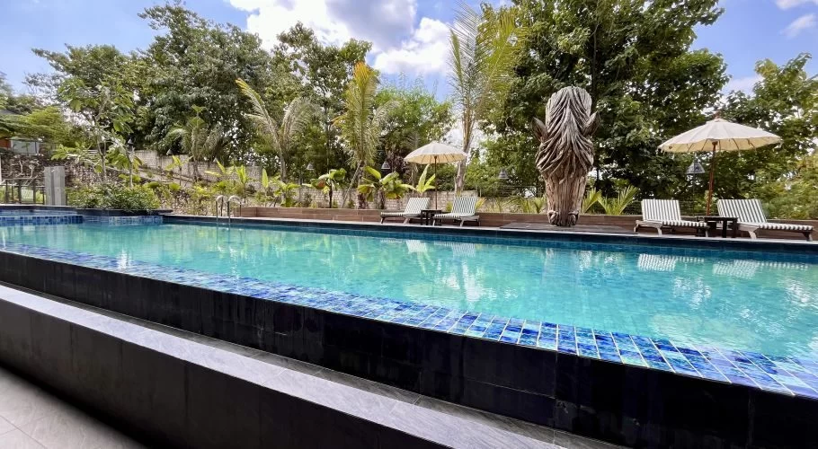 A long, blue tiled swimming pool is flanked by lush green trees, striped lounge chairs, and a large carved wooden horse head under a partly cloudy sky.