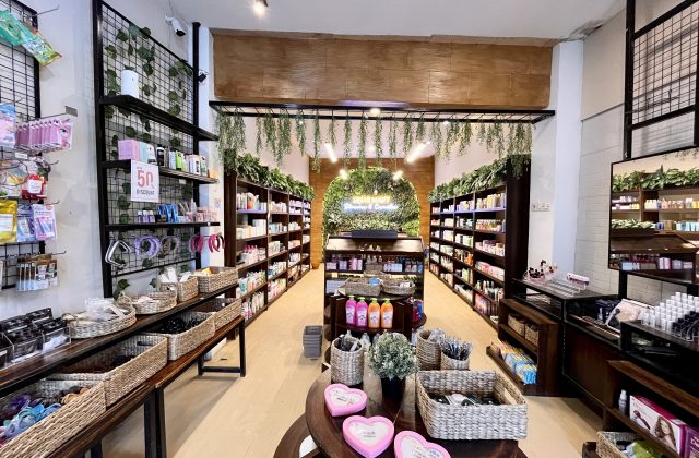 A brightly lit skincare store interior features dark wooden shelves packed with colorful products, wicker baskets, and a green wall with a neon sign at the back.
