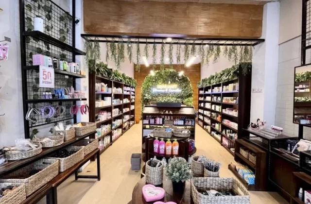 A brightly lit skincare store interior features dark wooden shelves packed with colorful products, wicker baskets, and a green wall with a neon sign at the back.