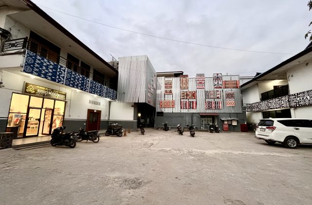 The exterior of Sinar Tambolaka Hotel features a paved parking lot, a souvenir shop, and buildings adorned with colorful Sumba ikat textiles under a cloudy sky.