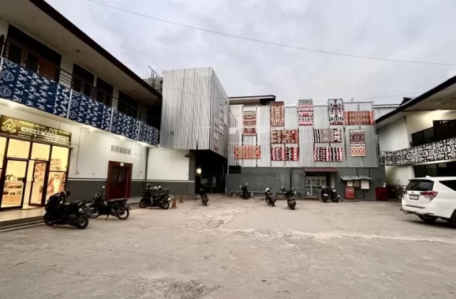 The exterior of Sinar Tambolaka Hotel features a paved parking lot, a souvenir shop, and buildings adorned with colorful Sumba ikat textiles under a cloudy sky.
