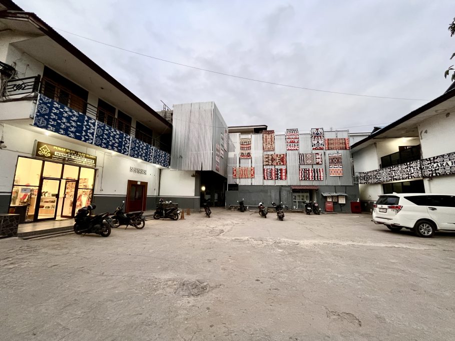 The exterior of Sinar Tambolaka Hotel features a paved parking lot, a souvenir shop, and buildings adorned with colorful Sumba ikat textiles under a cloudy sky.