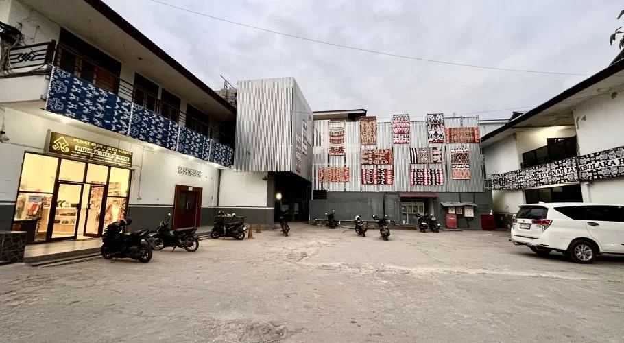The exterior of Sinar Tambolaka Hotel features a paved parking lot, a souvenir shop, and buildings adorned with colorful Sumba ikat textiles under a cloudy sky.