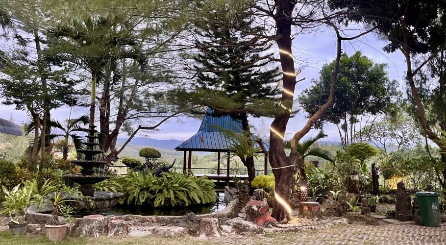 Lush green garden with a tiered stone fountain, a blue-roofed gazebo, and trees wrapped with string lights, set against distant mountains under a cloudy sky.
