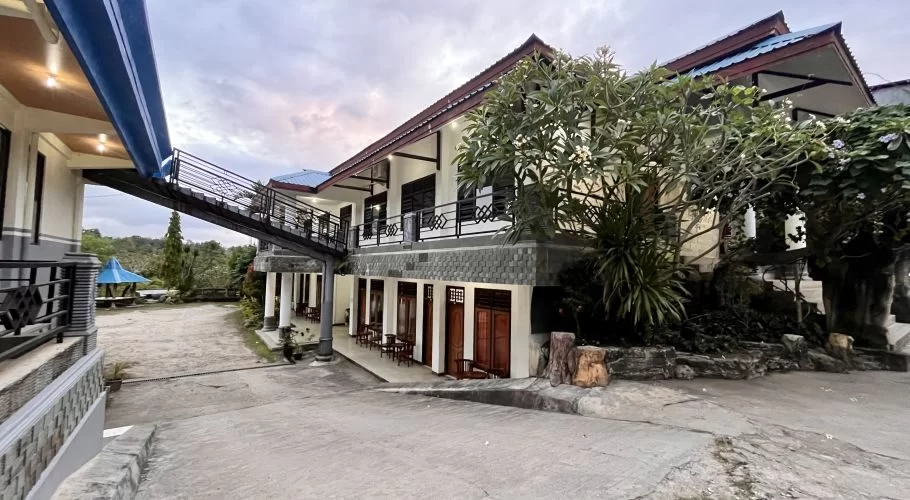 A white multi-story hotel building with dark wooden doors and a blue roof stands beside a paved driveway, surrounded by green trees and a distant blue gazebo under a cloudy sky.