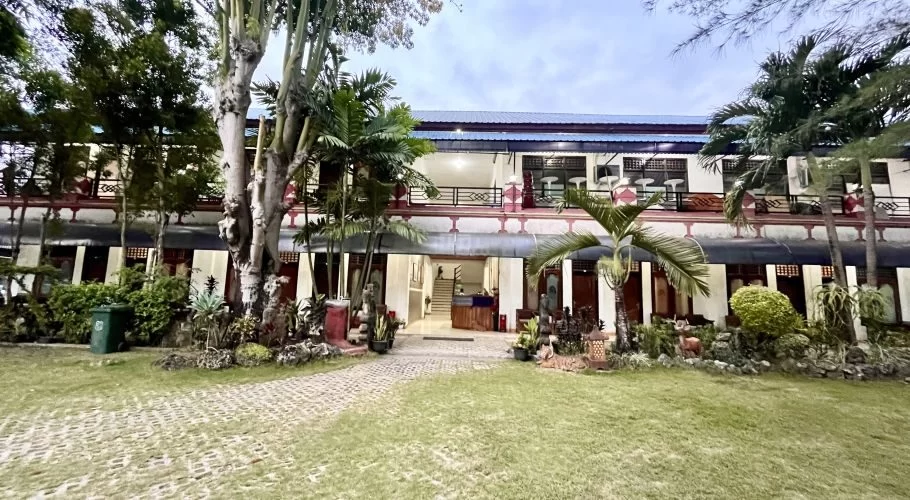The Sinar Tambolaka Hotel, a two-story white building with red accents, stands behind a lush green lawn and tropical trees under an overcast sky.