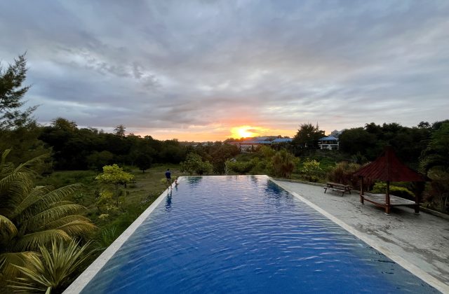 A blue infinity pool overlooks a lush green landscape and distant buildings under a cloudy sky with a vibrant orange sunset.
