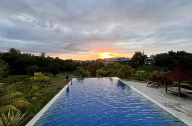 A blue infinity pool overlooks a lush green landscape and distant buildings under a cloudy sky with a vibrant orange sunset.