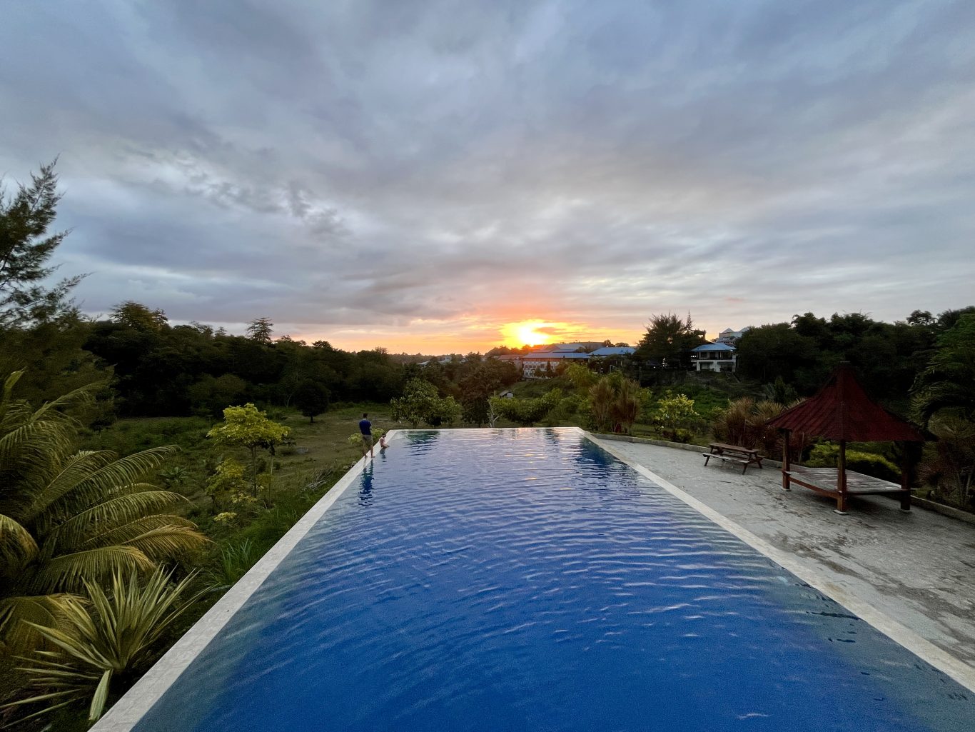 A blue infinity pool overlooks a lush green landscape and distant buildings under a cloudy sky with a vibrant orange sunset.