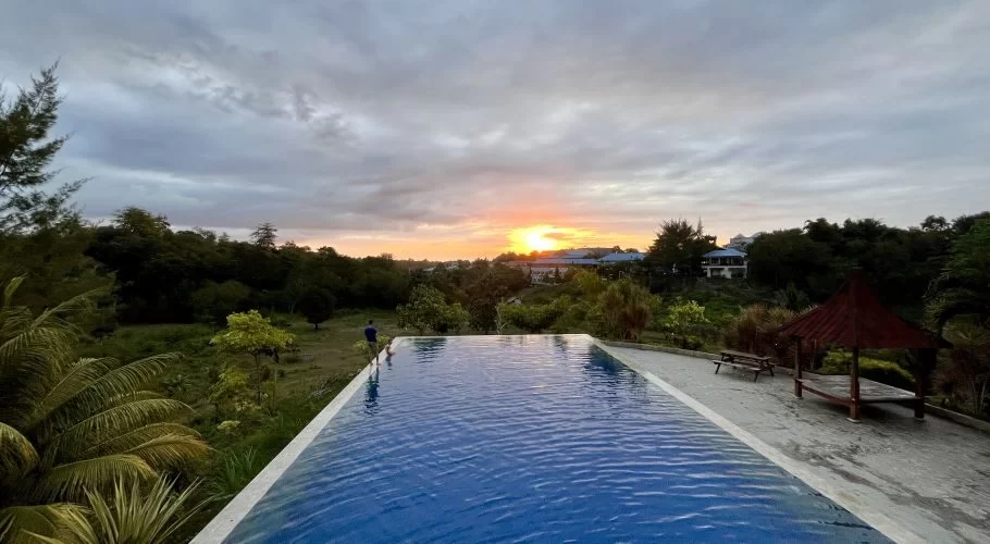 A blue infinity pool overlooks a lush green landscape and distant buildings under a cloudy sky with a vibrant orange sunset.