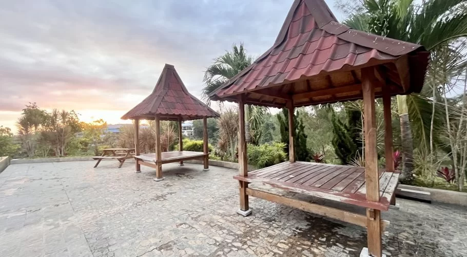 Two traditional wooden gazebos with red tiled roofs stand on a paved area amidst tropical plants, illuminated by a sunset glow under a cloudy sky.