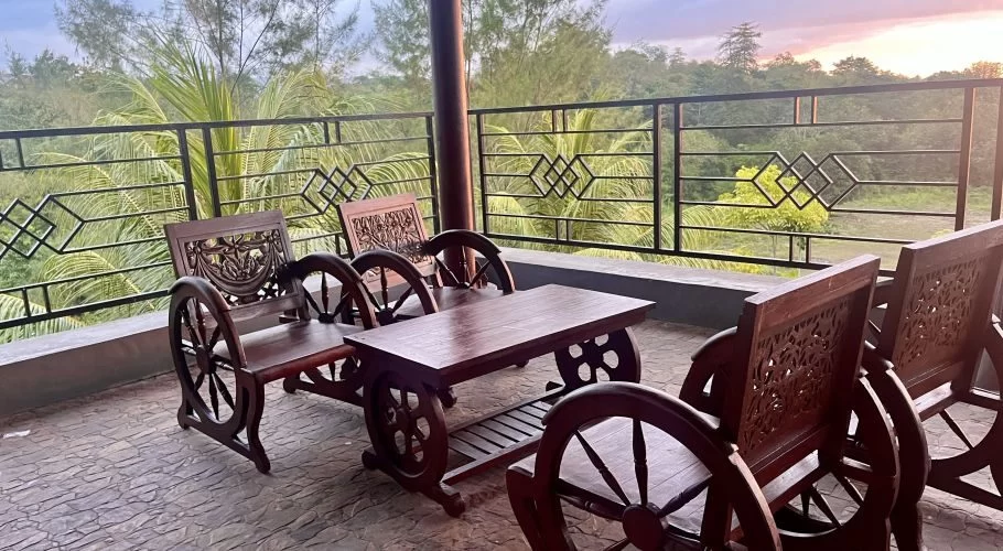 Dark wooden chairs and a table with carved details sit on a stone patio overlooking lush green trees and a warm-hued sunset sky.