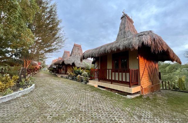 A row of traditional Sumba villas with tall thatched roofs lines a paved path, surrounded by green trees and grass under a cloudy sky.