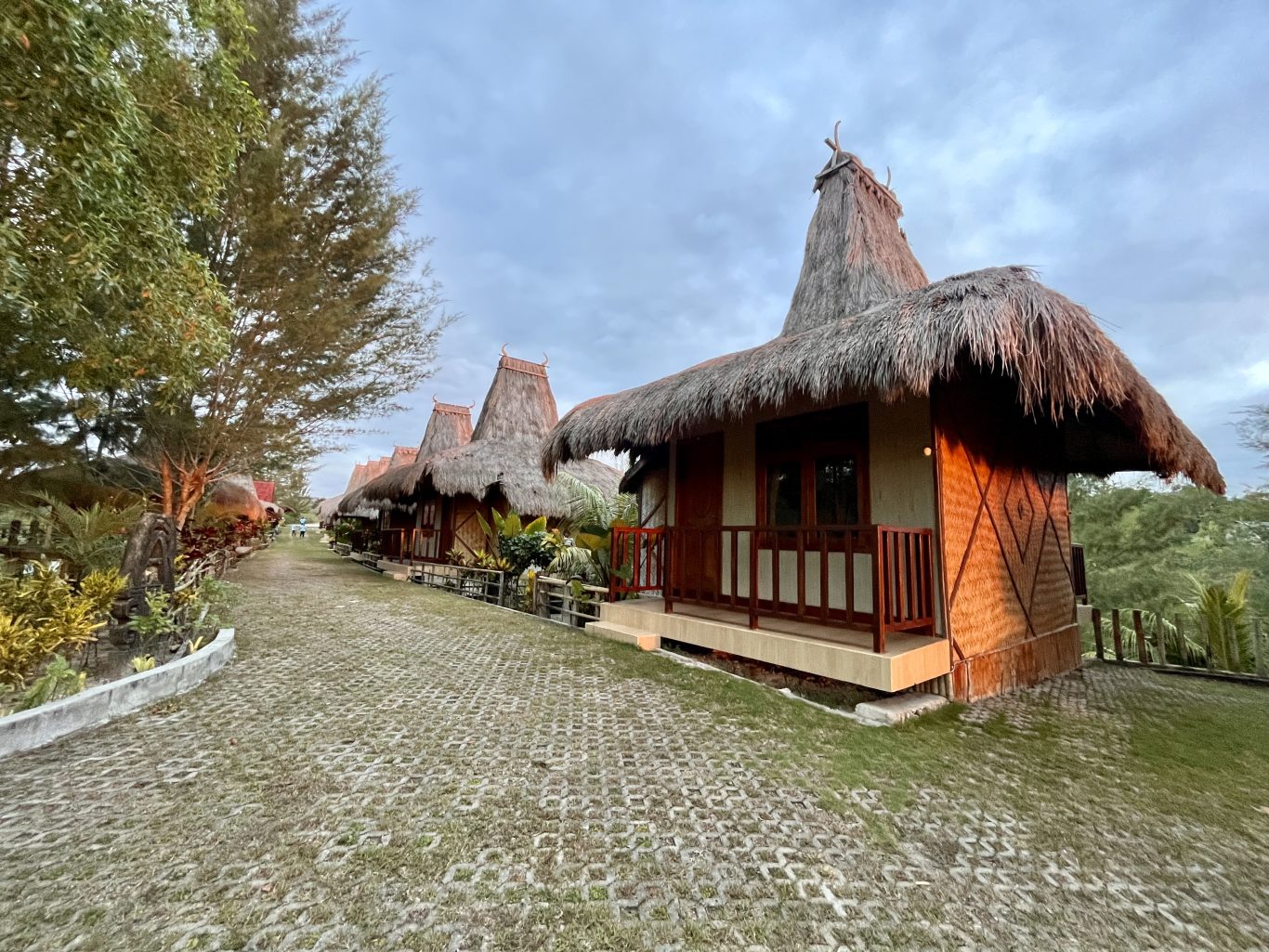 A row of traditional Sumba villas with tall thatched roofs lines a paved path, surrounded by green trees and grass under a cloudy sky.