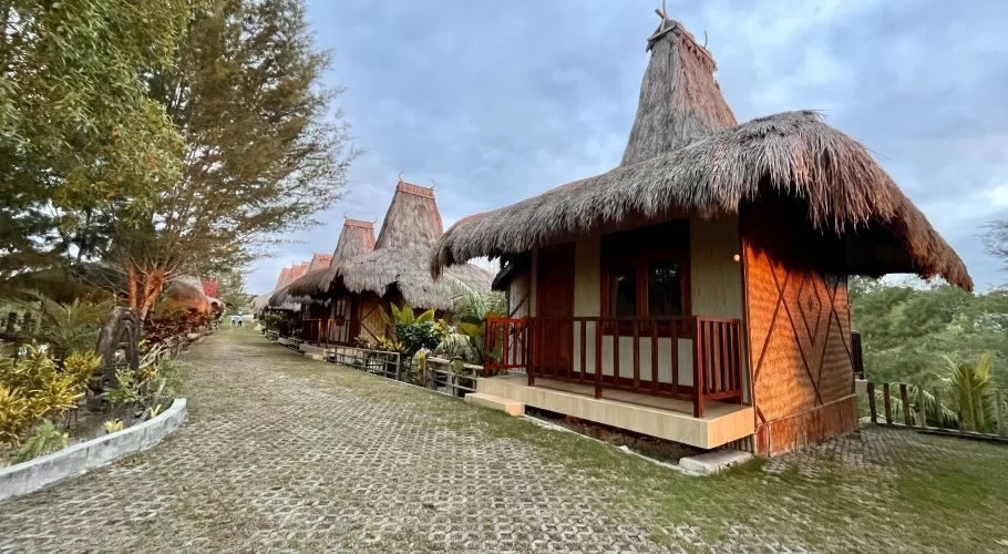 A row of traditional Sumba villas with tall thatched roofs lines a paved path, surrounded by green trees and grass under a cloudy sky.