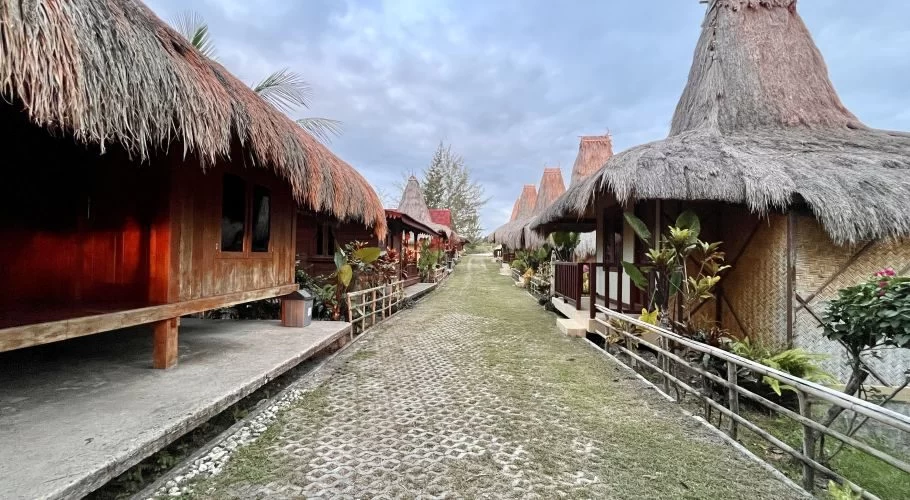 A paved pathway stretches between two rows of elevated wooden villas with traditional thatched roofs and tropical plants under an overcast sky.