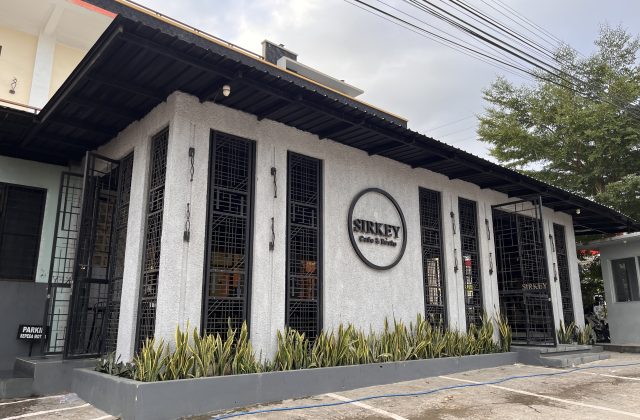 The modern white exterior of Sirkey Cafe & Bistro features black-framed windows, a dark roof, and green plants in a planter under a cloudy sky.