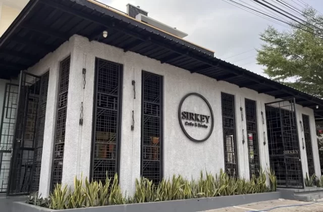The modern white exterior of Sirkey Cafe & Bistro features black-framed windows, a dark roof, and green plants in a planter under a cloudy sky.