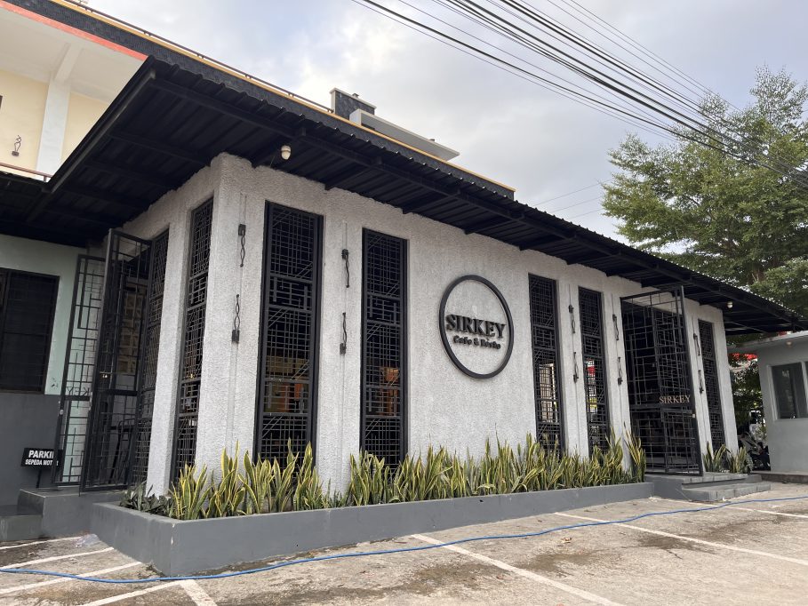 The modern white exterior of Sirkey Cafe & Bistro features black-framed windows, a dark roof, and green plants in a planter under a cloudy sky.