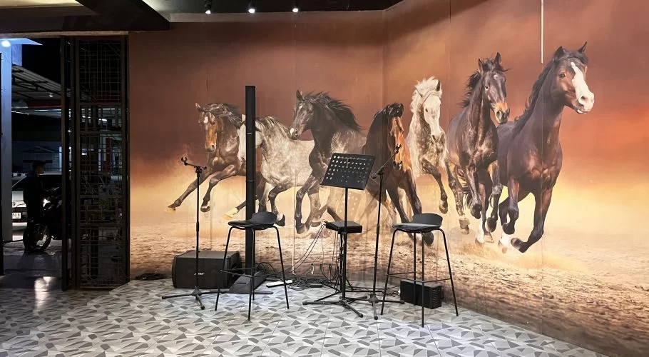 A stage with three black bar stools and a music stand is set against a large mural of galloping horses in a warm-toned landscape.