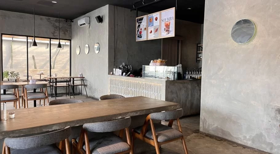 A modern cafe interior with concrete walls, black ceilings, wooden tables, grey chairs, and a counter displaying menu boards, located in Waikabukak.
