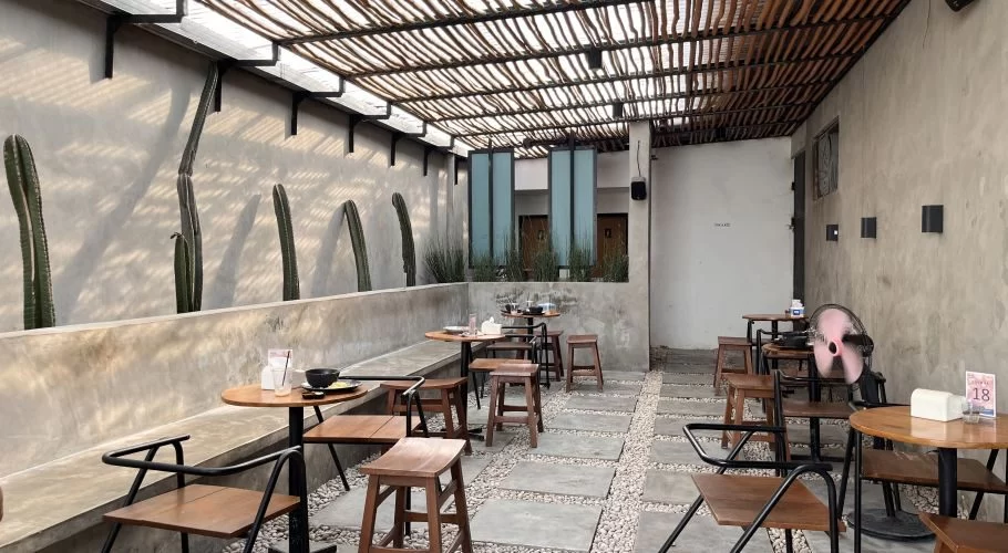 Outdoor cafe seating features concrete walls with tall cacti, wooden tables and stools on a gravel and paver floor, under a slatted roof.