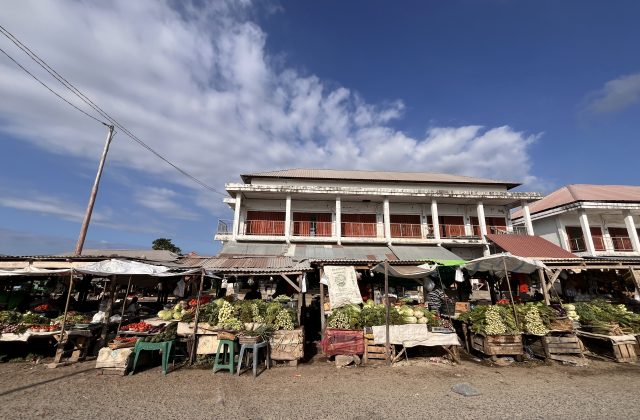 A bustling traditional market in Sumba features numerous stalls overflowing with fresh green vegetables under a bright blue sky, with simple buildings in the background.
