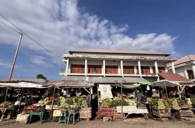 A bustling traditional market in Sumba features numerous stalls overflowing with fresh green vegetables under a bright blue sky, with simple buildings in the background.