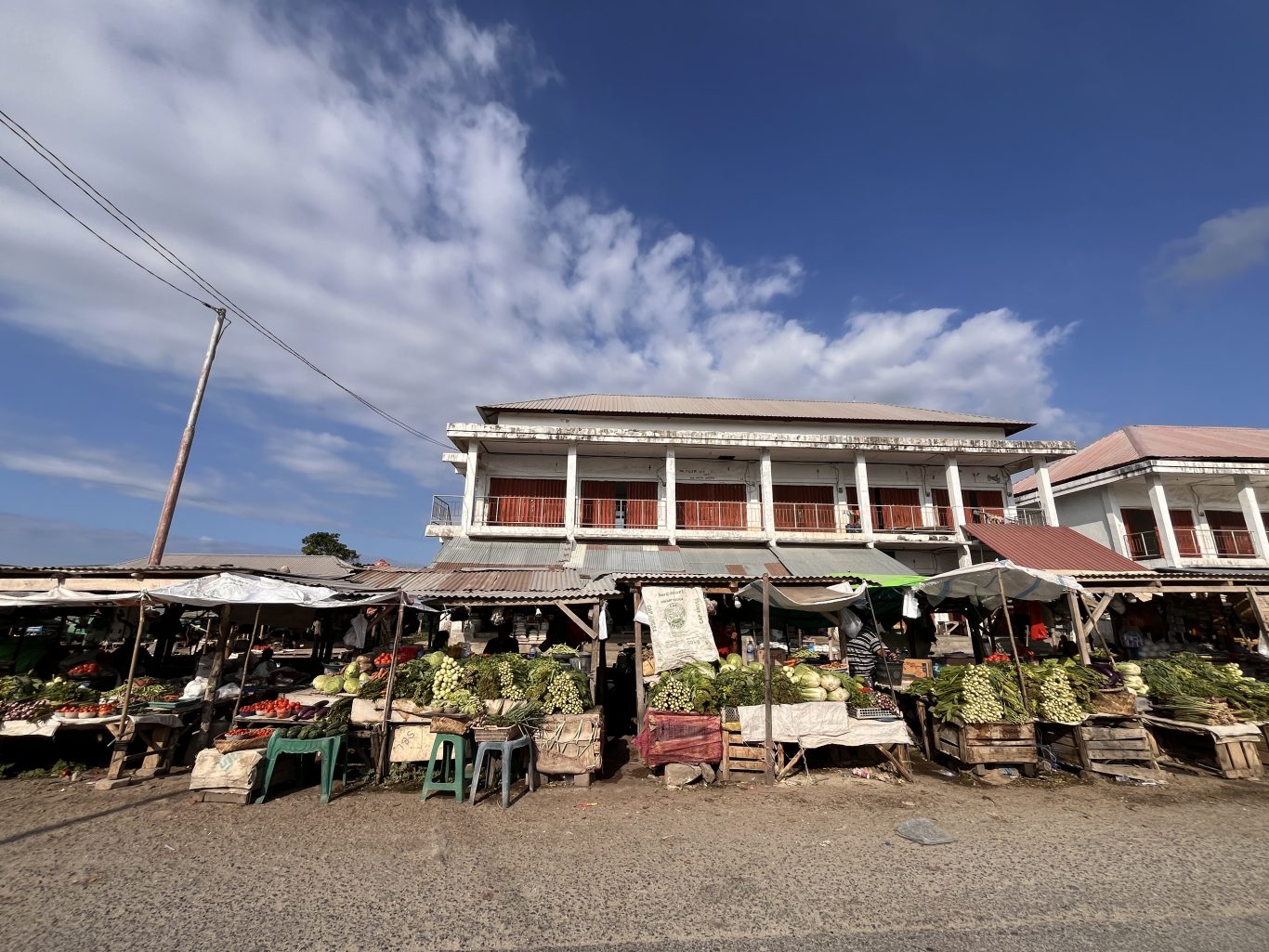A bustling traditional market in Sumba features numerous stalls overflowing with fresh green vegetables under a bright blue sky, with simple buildings in the background.