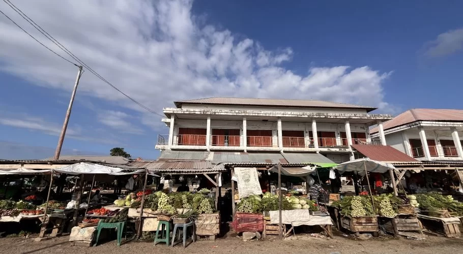 A bustling traditional market in Sumba features numerous stalls overflowing with fresh green vegetables under a bright blue sky, with simple buildings in the background.