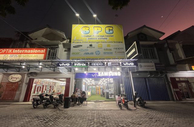 A brightly lit SPC electronics store with a large yellow sign stands out on a street at dusk, with people sitting outside and motorcycles parked.