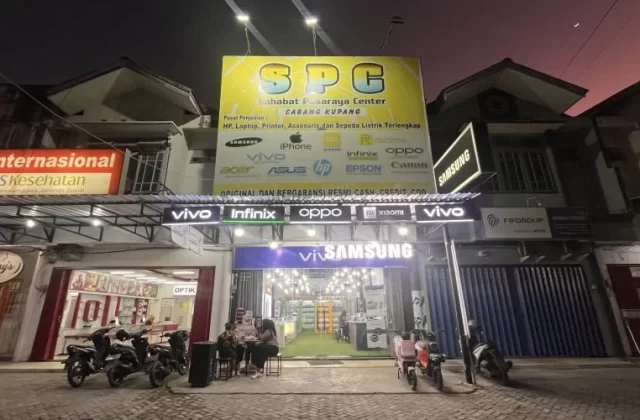A brightly lit SPC electronics store with a large yellow sign stands out on a street at dusk, with people sitting outside and motorcycles parked.