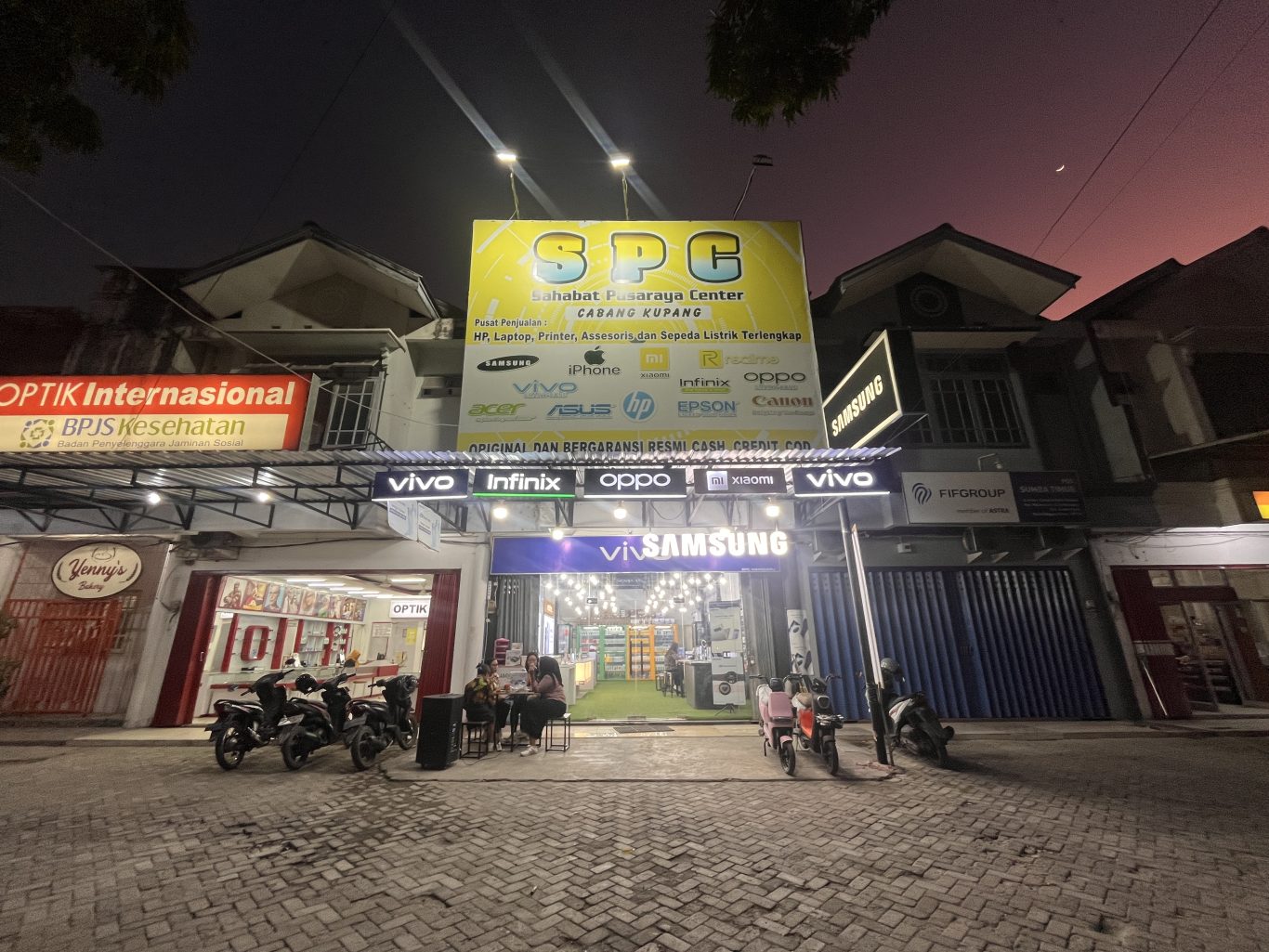 A brightly lit SPC electronics store with a large yellow sign stands out on a street at dusk, with people sitting outside and motorcycles parked.