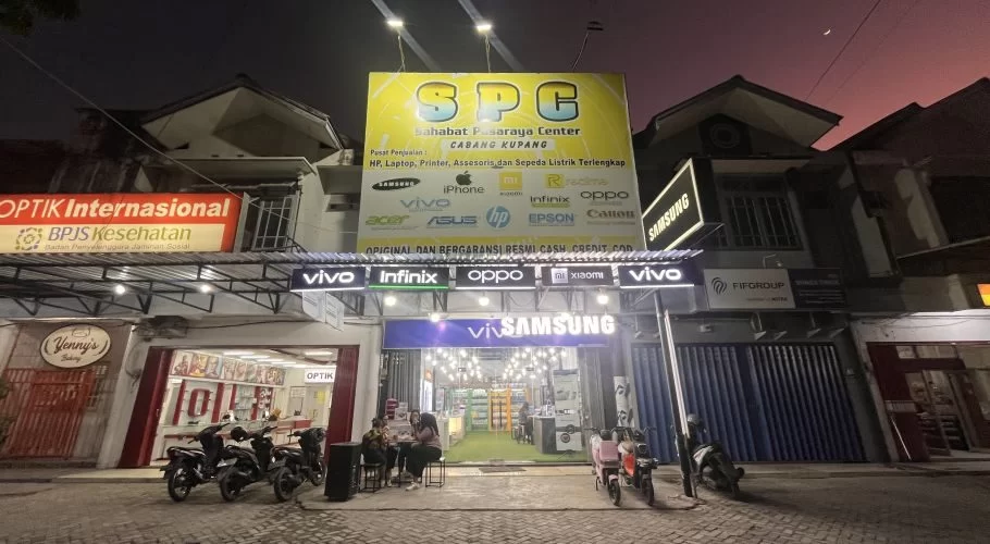 A brightly lit SPC electronics store with a large yellow sign stands out on a street at dusk, with people sitting outside and motorcycles parked.