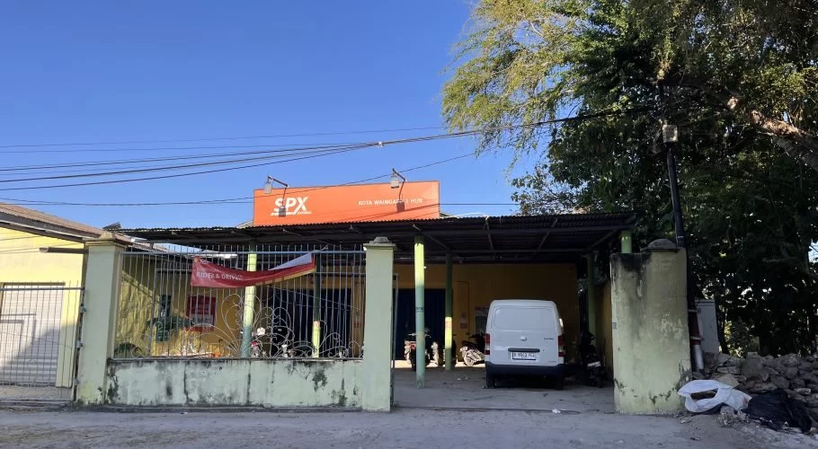 A yellow SPX Express Waingapu Hub building with an orange sign, a white delivery van, and motorcycles parked under a canopy on a clear day.