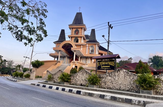 The orange St. Petrus & Paulus Catholic church in Waikabubak features multiple dark spires, statues, and a stone wall along a road under a clear sky.