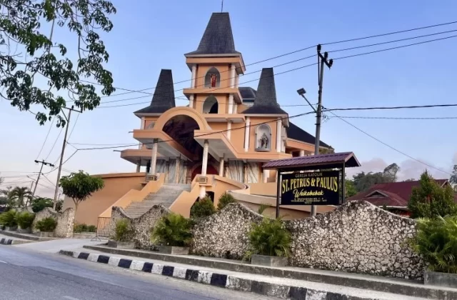 The orange St. Petrus & Paulus Catholic church in Waikabubak features multiple dark spires, statues, and a stone wall along a road under a clear sky.