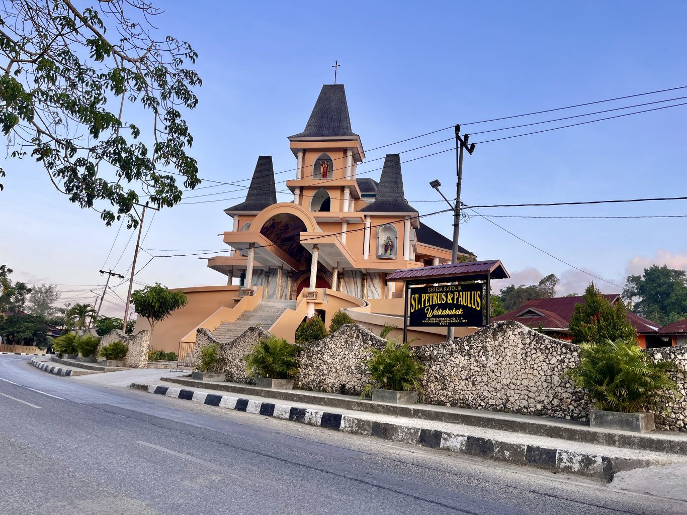 The orange St. Petrus & Paulus Catholic church in Waikabubak features multiple dark spires, statues, and a stone wall along a road under a clear sky.