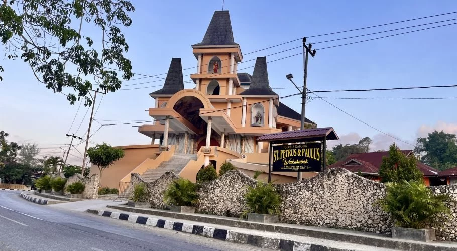 The orange St. Petrus & Paulus Catholic church in Waikabubak features multiple dark spires, statues, and a stone wall along a road under a clear sky.