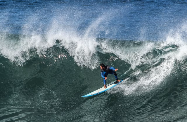 A surfer in a blue wetsuit rides a large, powerful right-hand ocean wave, creating white spray against the deep blue and green water.
