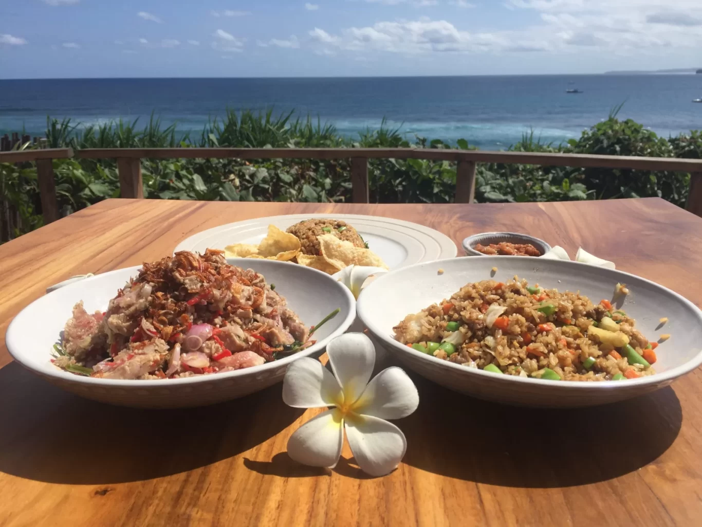 Two bowls of colorful Indonesian food and a white frangipani flower sit on a wooden table overlooking a deep blue ocean and green foliage under a bright sky.