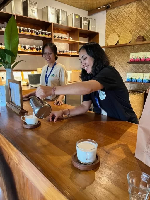 A smiling woman with dark hair pours a light-colored drink from a silver pitcher into a clear mug on a wooden counter, while another woman smiles behind her. Shelves with various products line the background.