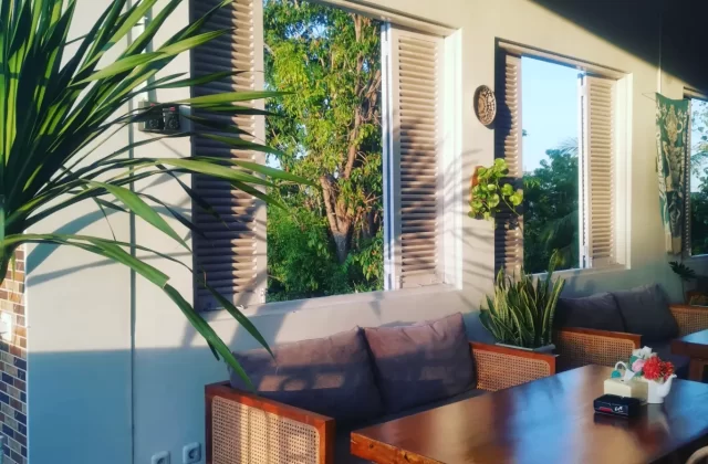 A sunlit outdoor cafe features a long wooden table, rattan sofas with grey cushions, and lush green plants, with open windows revealing vibrant trees.