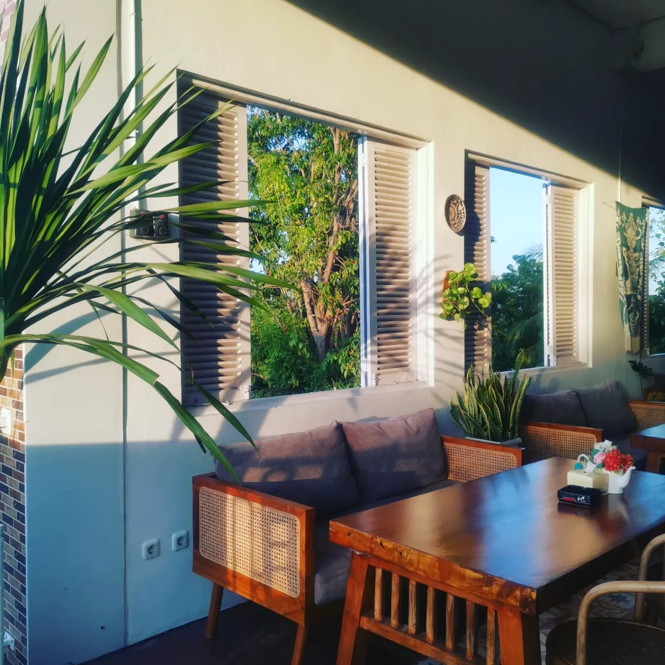 A sunlit outdoor cafe features a long wooden table, rattan sofas with grey cushions, and lush green plants, with open windows revealing vibrant trees.