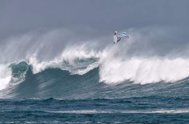A windsurfer is airborne above a massive, breaking ocean wave under a dark gray sky, with white spray erupting from the wave.
