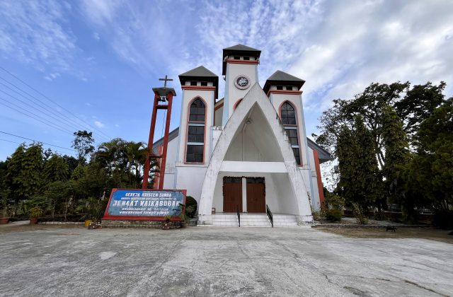 A white church with red trim and three spires stands under a blue sky, surrounded by lush green trees and a sign in front.