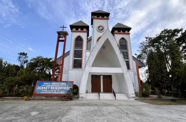 A white church with red trim and three spires stands under a blue sky, surrounded by lush green trees and a sign in front.